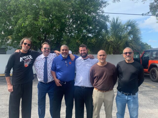 Six men standing together outdoors, smiling.