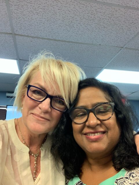 Two women smiling, wearing glasses, indoors.